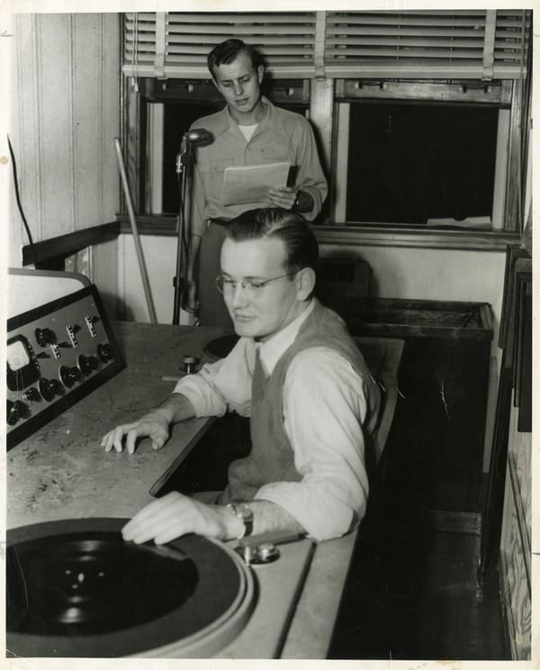 A person sitting in front of a turntable while another person stands at a microphone with windows behind him.