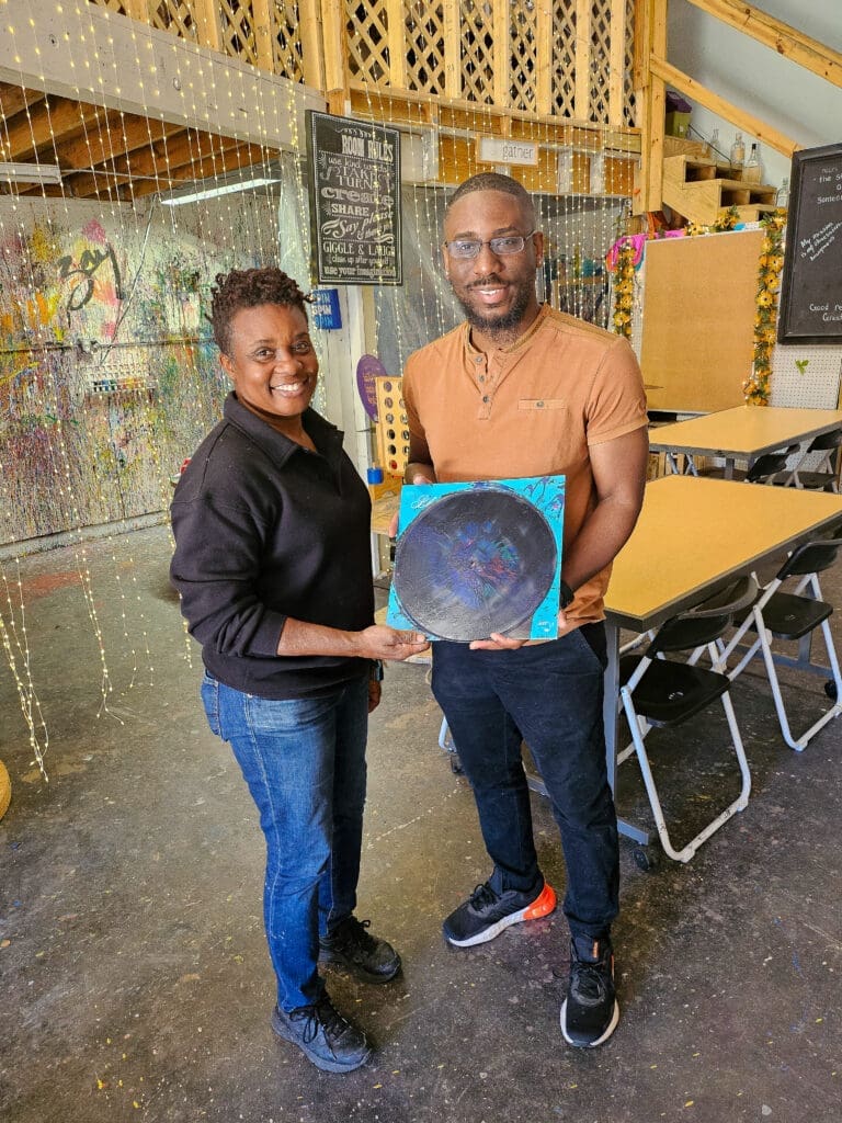 Woman and man stand next to each other. Man holds a record with paint on it.