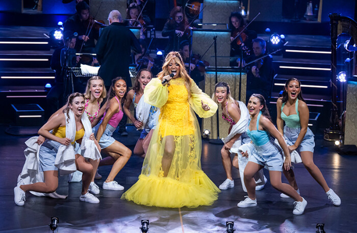 woman in yellow singing, surrounded by dancers image