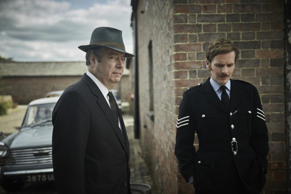 Two men dressed in old fashion clothing near a brick wall.