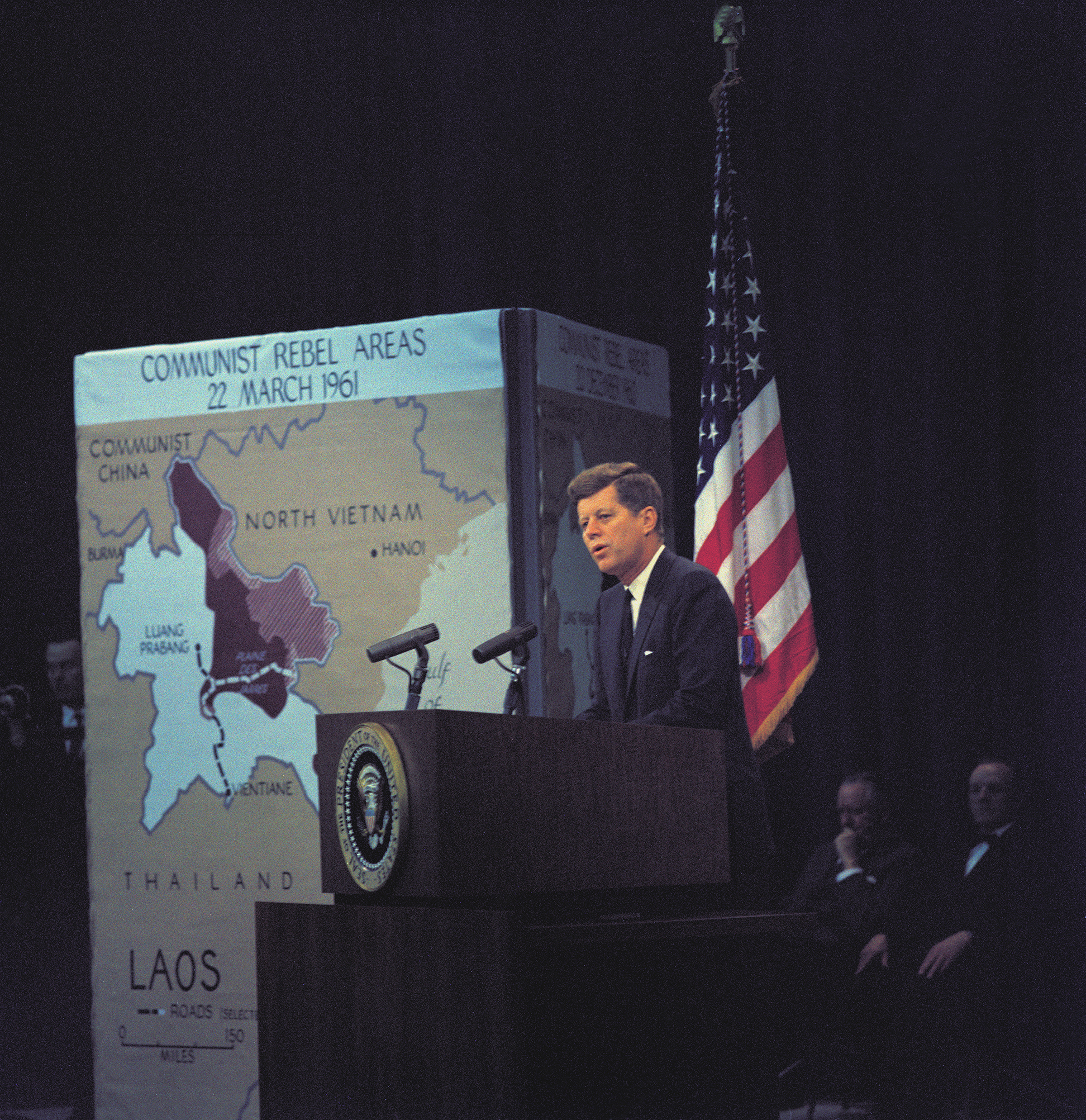 John F Kennedy giving a speech with a map of Laos on display image