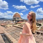 Chloe is shown in a pink dress on top of a roof in urban italy.