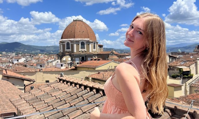 Chloe is shown in a pink dress on top of a roof in urban italy.