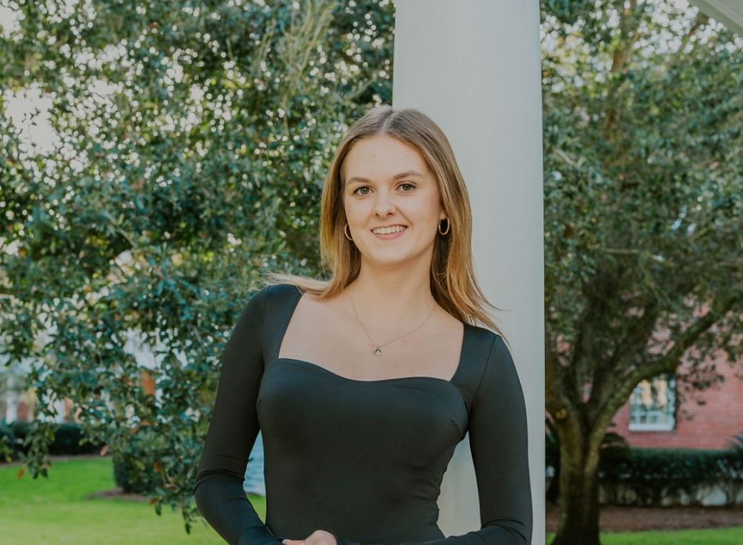 Hannah is pictured in a black dress in front of a white post and a large tree.