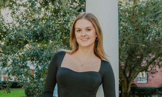 Hannah is pictured in a black dress in front of a white post and a large tree.