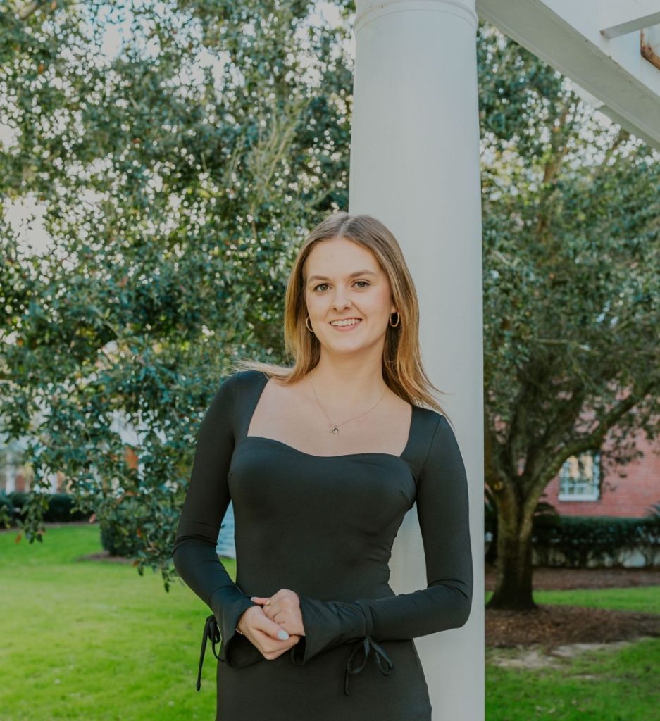 Hannah is pictured in a black dress in front of a white post and a large tree.