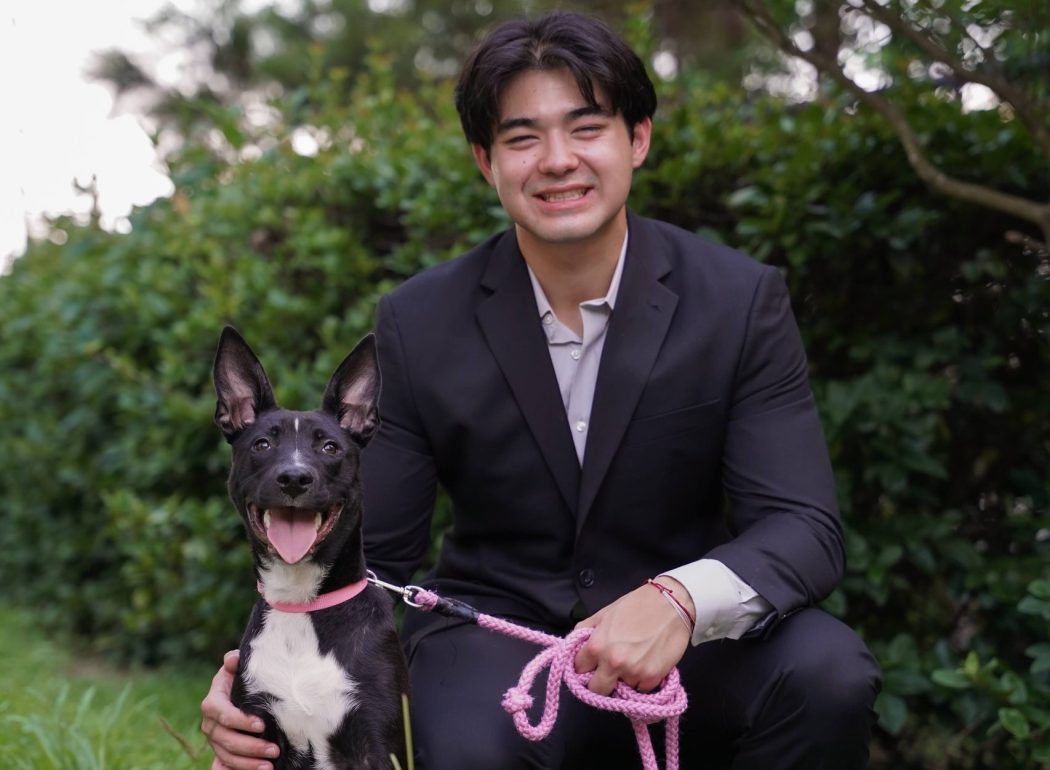 Kai Smith squats in a black suit on the grass in front of a push along with his dog.