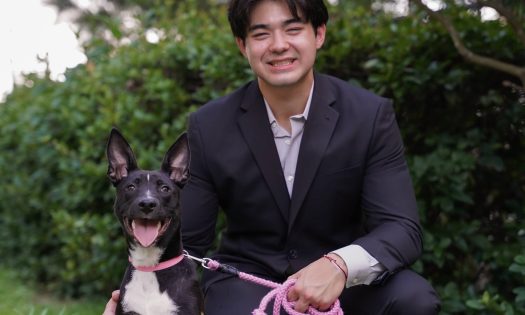 Kai Smith squats in a black suit on the grass in front of a push along with his dog.