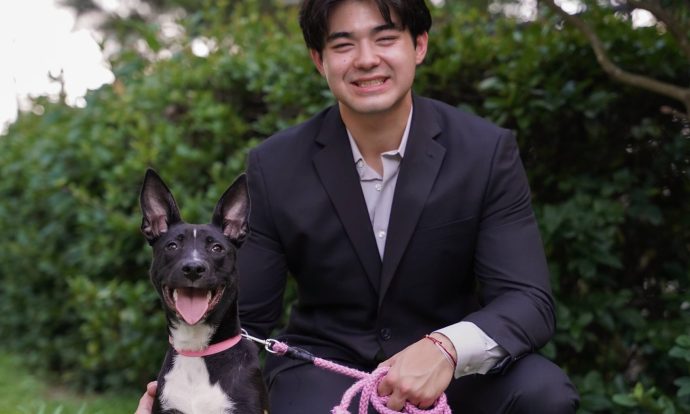 Kai Smith squats in a black suit on the grass in front of a push along with his dog.