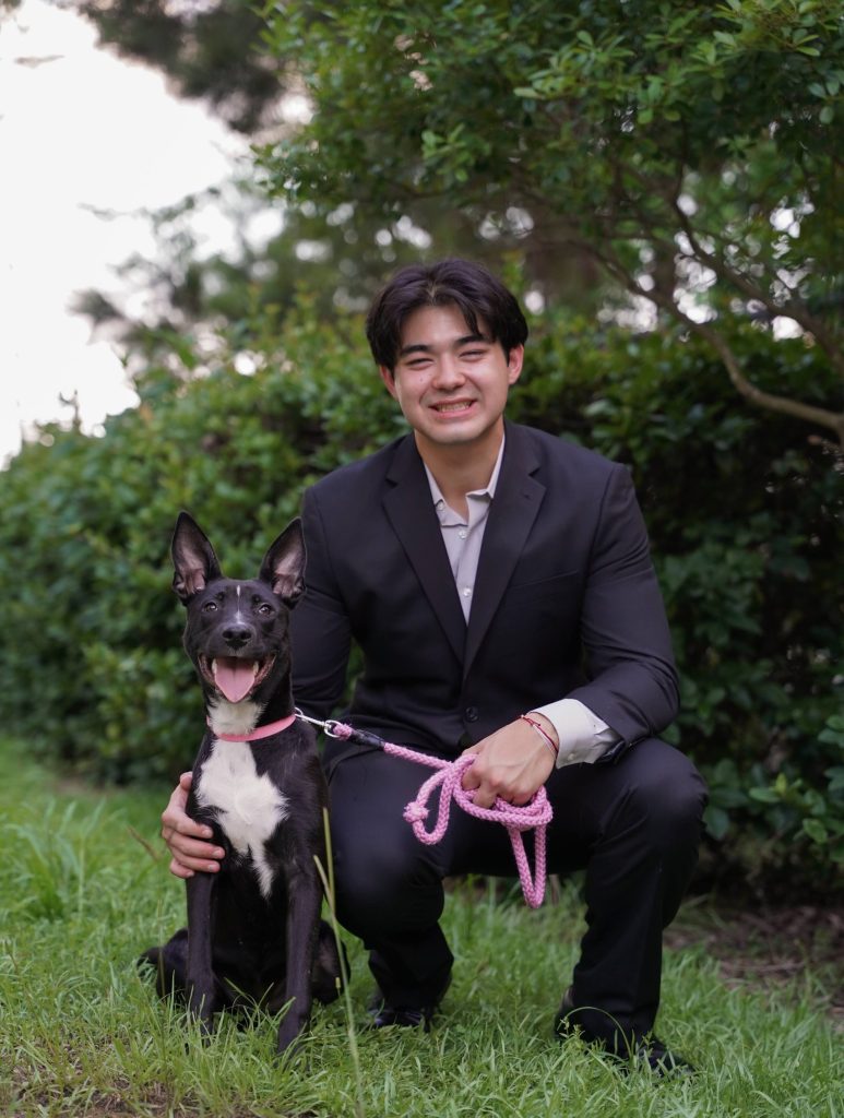 Kai Smith squats in a black suit on the grass in front of a push along with his dog.