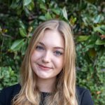 A smiling woman dressed in black stands in front of shrubbery.