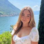 A smiling woman dressed white stands in front shrubbery, a lake and a mountain.