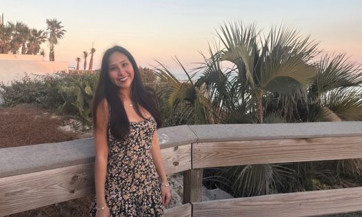 A smiling woman in a sundress stands on a wooden deck on a beach.