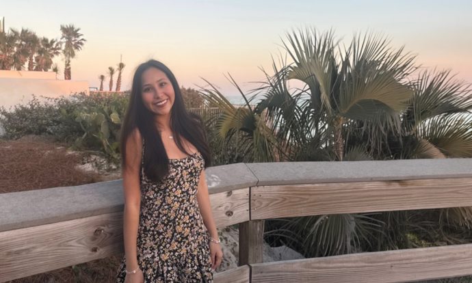 A smiling woman in a sundress stands on a wooden deck on a beach.
