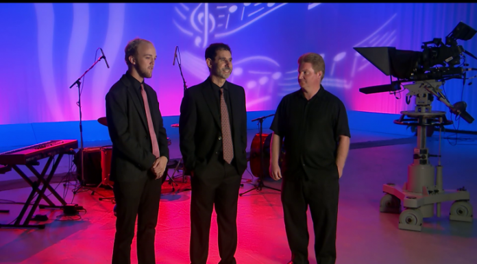 The Men and Music of the World Music Trio Three musicians stand on stage in pink and blue lit up room