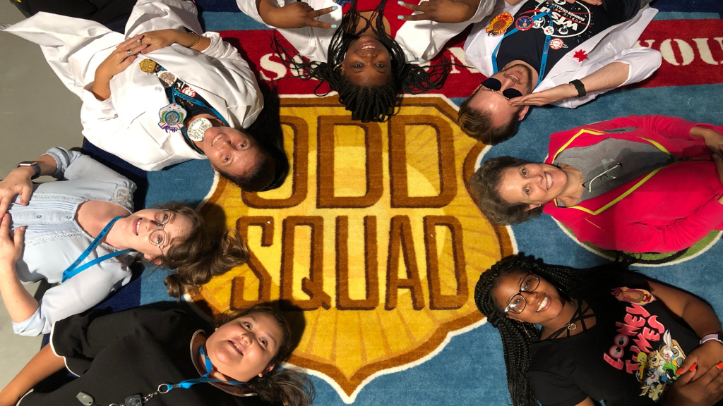 A group of people laying down in a circle around a carpet that says "Odd Squad"