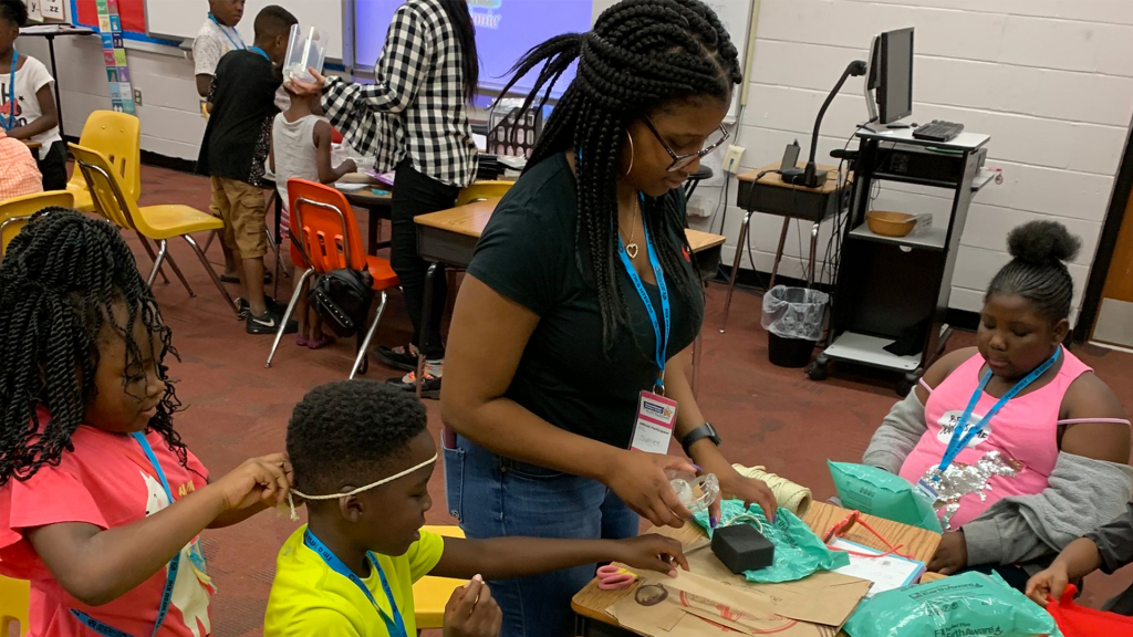 A young woman in black shirt and jeans  surrounded by echildren working on craft projects.