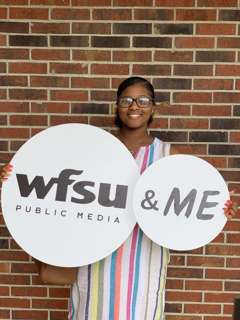 A young woman holding a sign wth two circles. One says "WFSU Public Media" and the other says "& Me" She is standing in front of a brick wall at WFSU