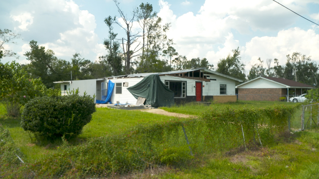 A small house with a severely damaged roof and tarps hanging off structure in a grassy yard.