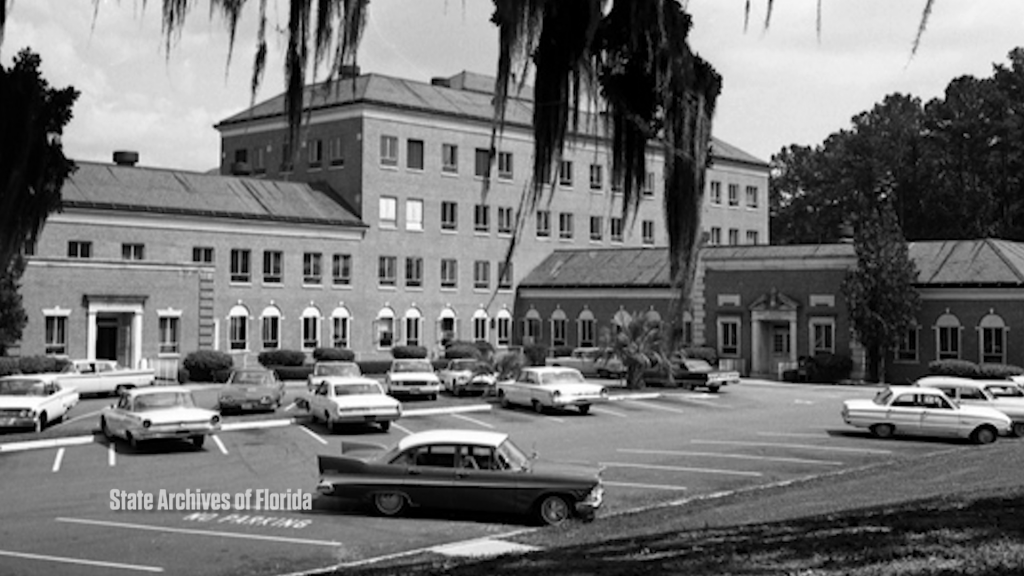 black and white photo of large brick building with cars from the 1950's parked in front.