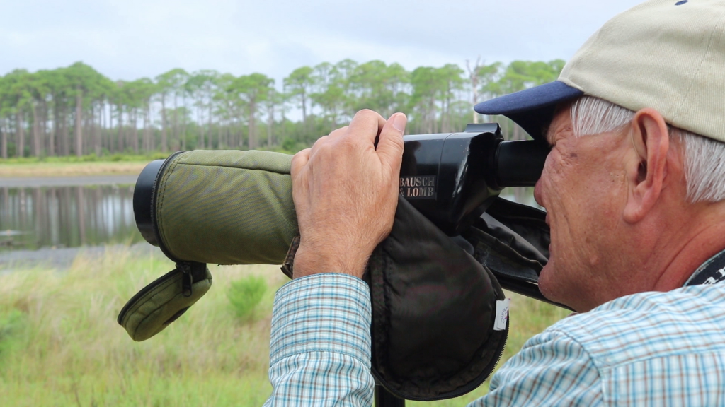 A man looking through a large viewfinder at trees. He is wearing a tan baseball cap and a plaid shirt.