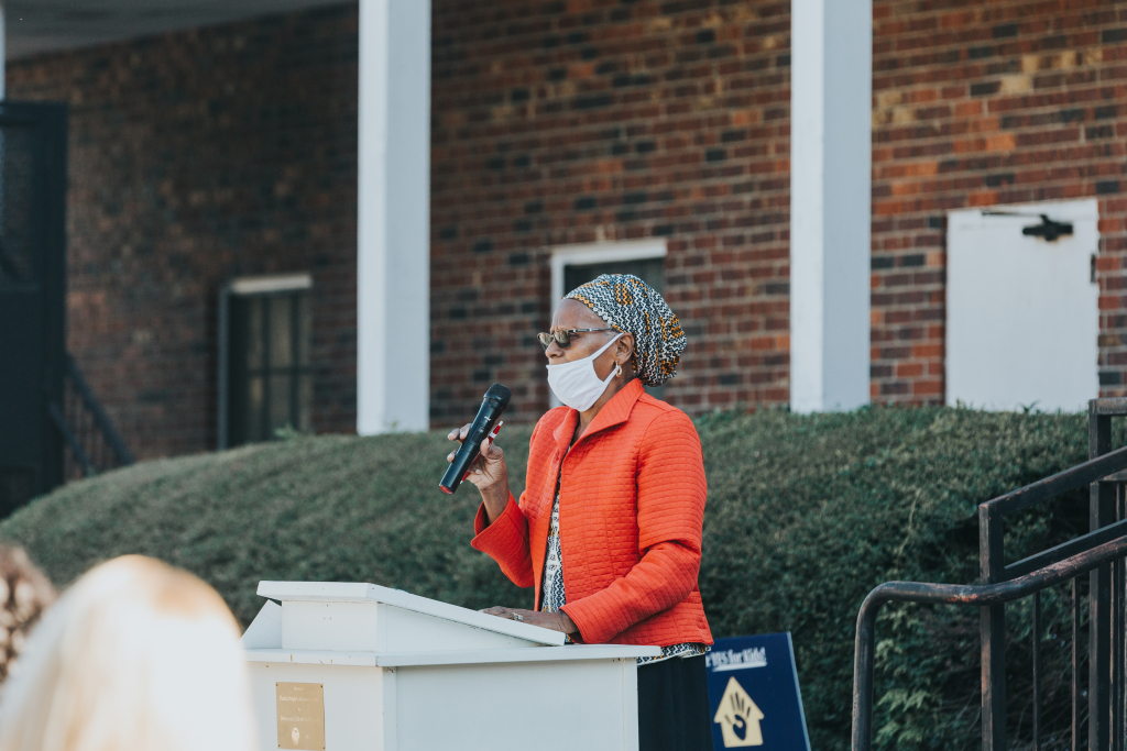 Woman in orange jacket holding microphone in front of brick building.
