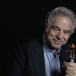 Itzhak Perlman wearing a suit and tie holding a cell phone