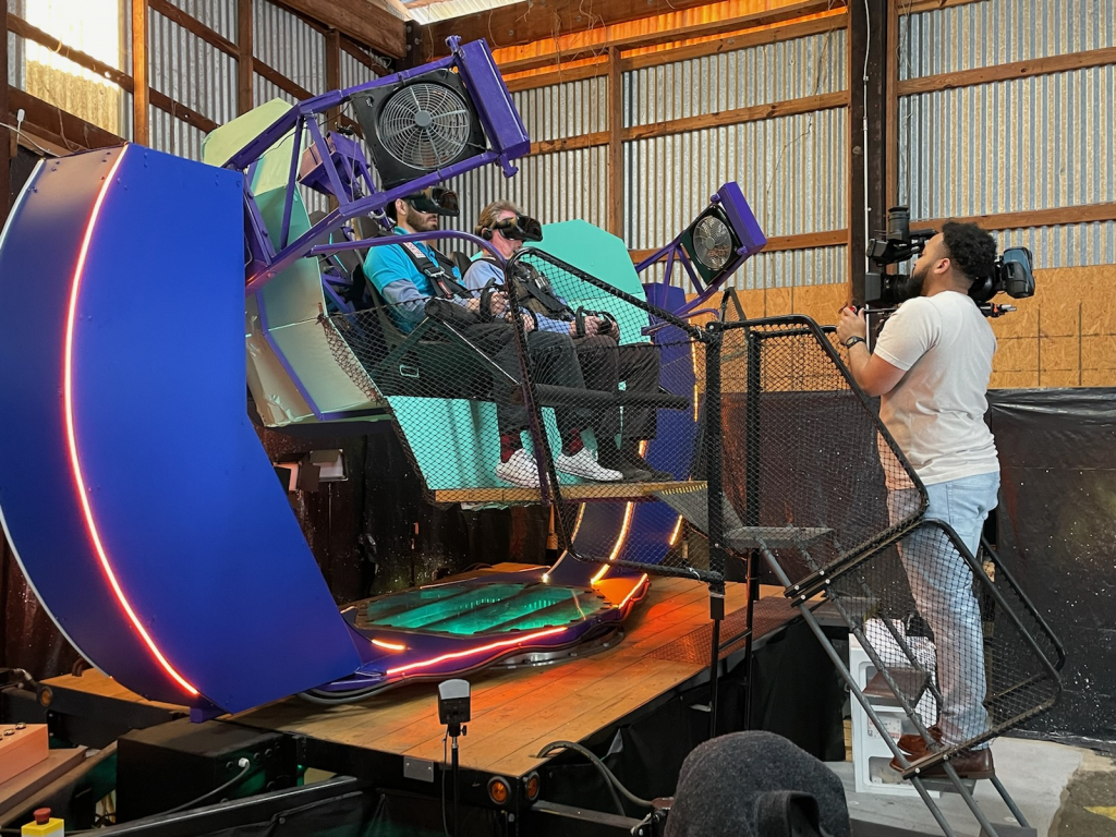 A man pointing a video camera at two other men seated on a motion platform. 