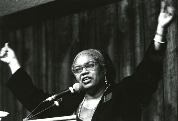 A woman with glasses and headband at podium holding arms up in air.