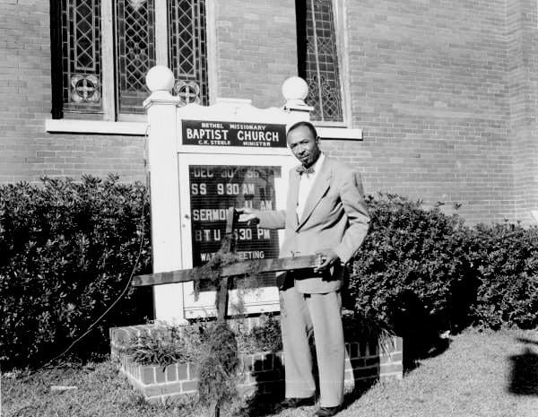 Man in suit standing in front of a church sign holding a burned cross