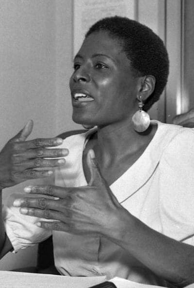 A black woman sitting at a table with papers in front of her talking to someone off camera