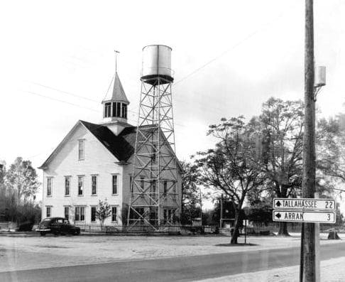 A black and white photo of a hold building with cupula and a water tower in front of it.  Signs with millage to Tallahassee and and Arran can be seen
