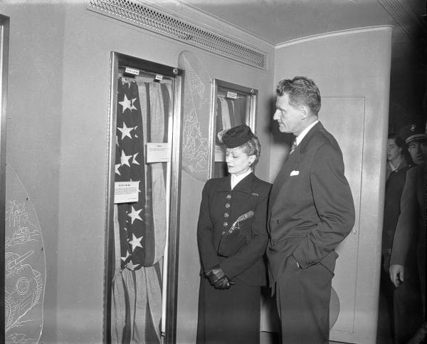 a woman in black stands next to a tall man in a suit. Both are looking at an American flag in a glass case.