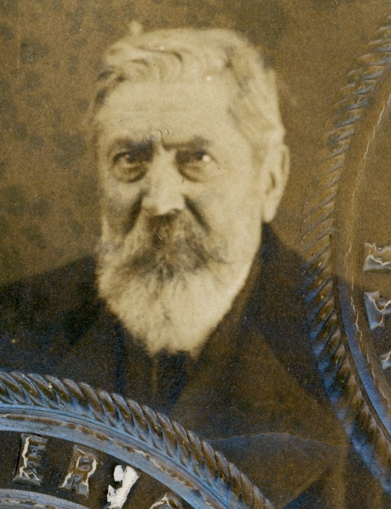 A man with white hair and a moustache and beard in black coat looking a photo in a sepia colored photo.