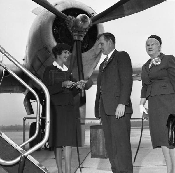 A flight attendant welcomes a well dressed man and a woman as they get ready to board a plane. The propeller can be seen behind them.