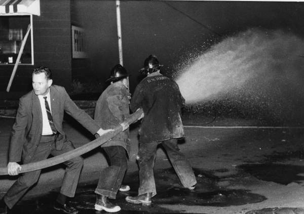 Black and white photo of two firefighters and a man in a suit and tie hold a hose to spray water on a building .
