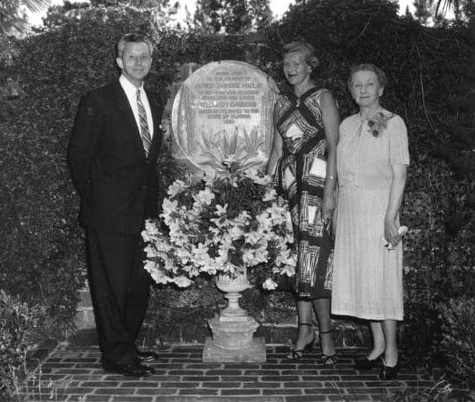 Governor LeRoy Collins in a suit and tie with two women in dresses with a planter in front of them with flowers.  Behind them is a medallion with writing.
