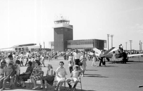 A group of people sitting and standing in front of an airplane and an airport control tower.