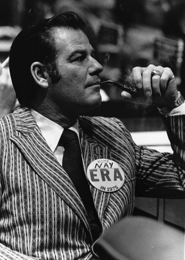 A man in a striped suit jacket and black tie smokes a pipe while wearing a large button that says 