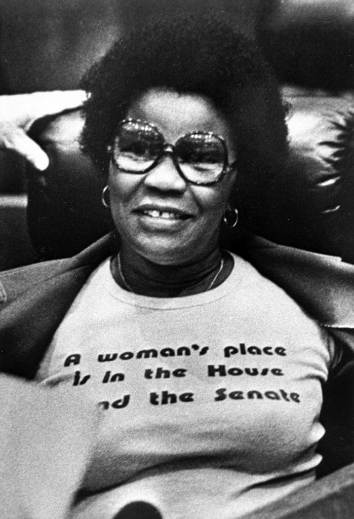 A woman wearing glasses wearing a shirt that says A woman's place is in the House and the Senate.