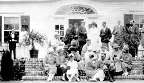 a black and white photo of a group of people standing and sitting on the brick stairs and porch of a white building in front of a open door.