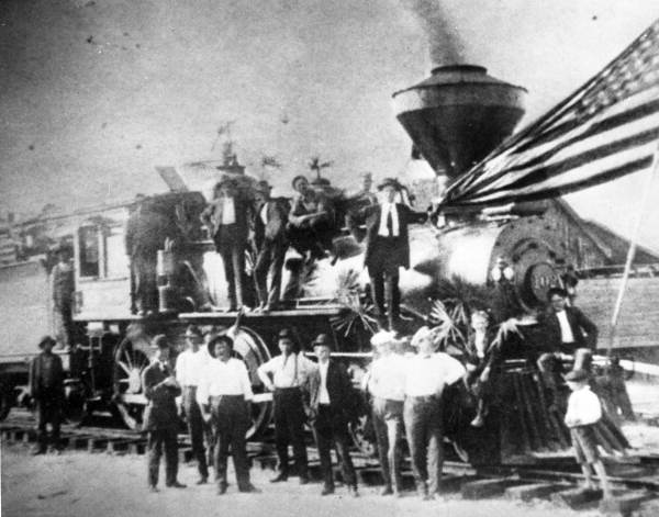 A vintage photo of a group of people standing in front of and on a locomotive.  An American flag can be seen in the front of the train.