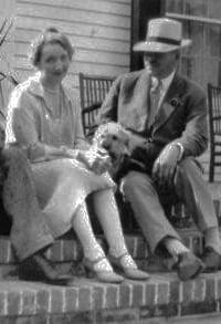 A woman in a dress, a dog and a man in a suit and hat sitting on brick steps of a porch