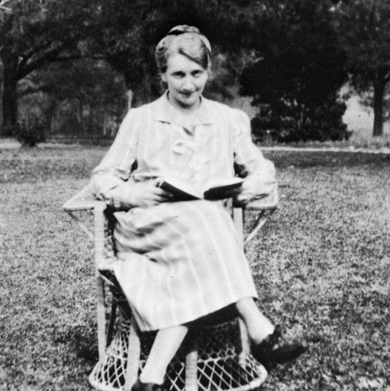 A woman in a stripped dress sitting in a chair with an open book on her lap.  She is looking up at the camera.