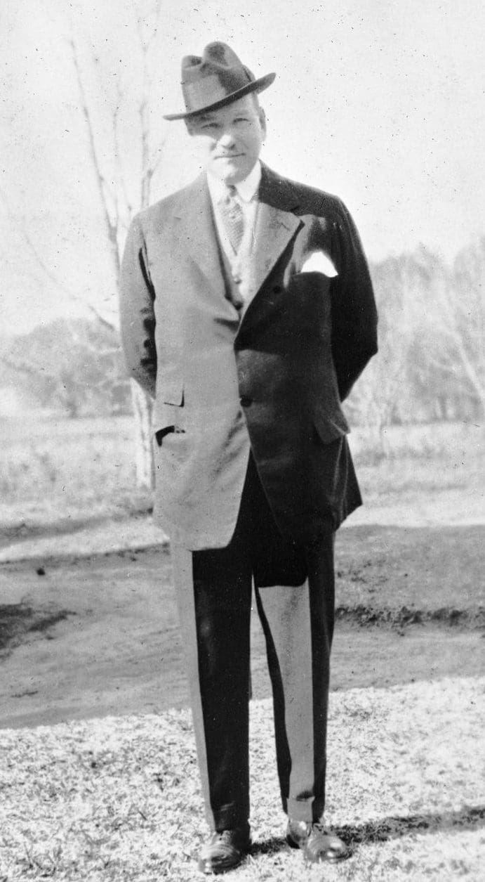 A man with moustache  standing in a field with a suit, tie and hat