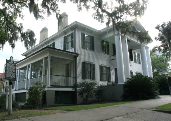 A  white house with black trim.  There are columns, a side porch, a historical marker, a sidewalk and trees in the background and branches overhead.