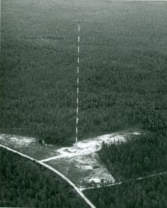 TV transmitter tower stands tall above a forest next to road.,