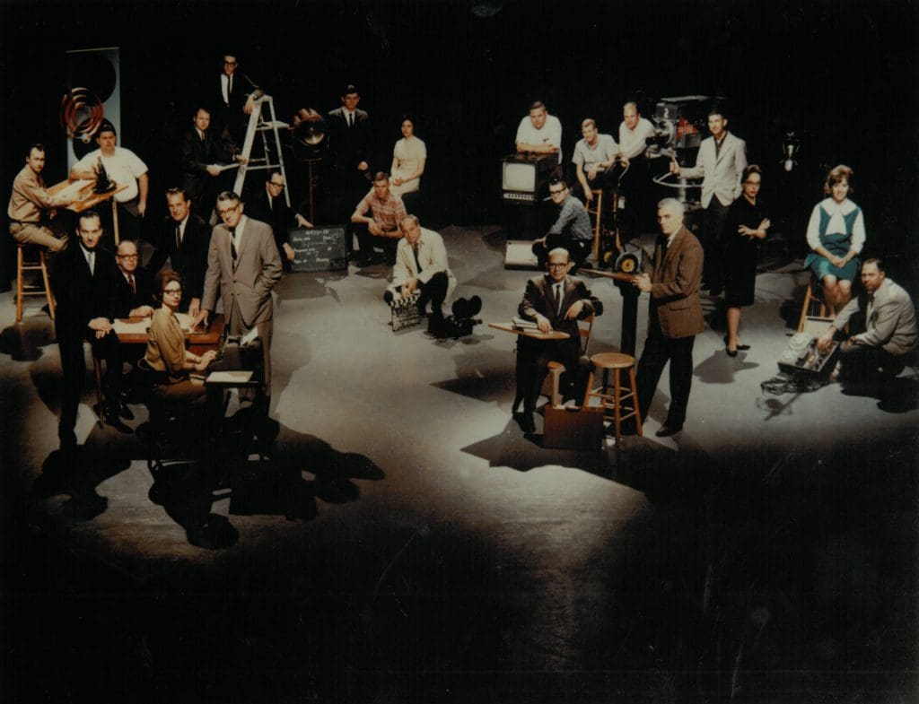 group of people standing in groups around television and office equipment in a large space with shadow