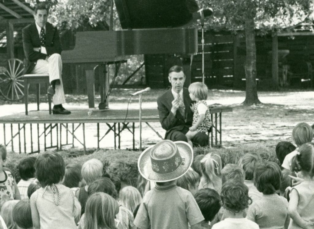 Fred Rogers sits in front of a stage with a piano where another man sits.  A child sits on Fred Roger's lap as many children sit in front of him. 