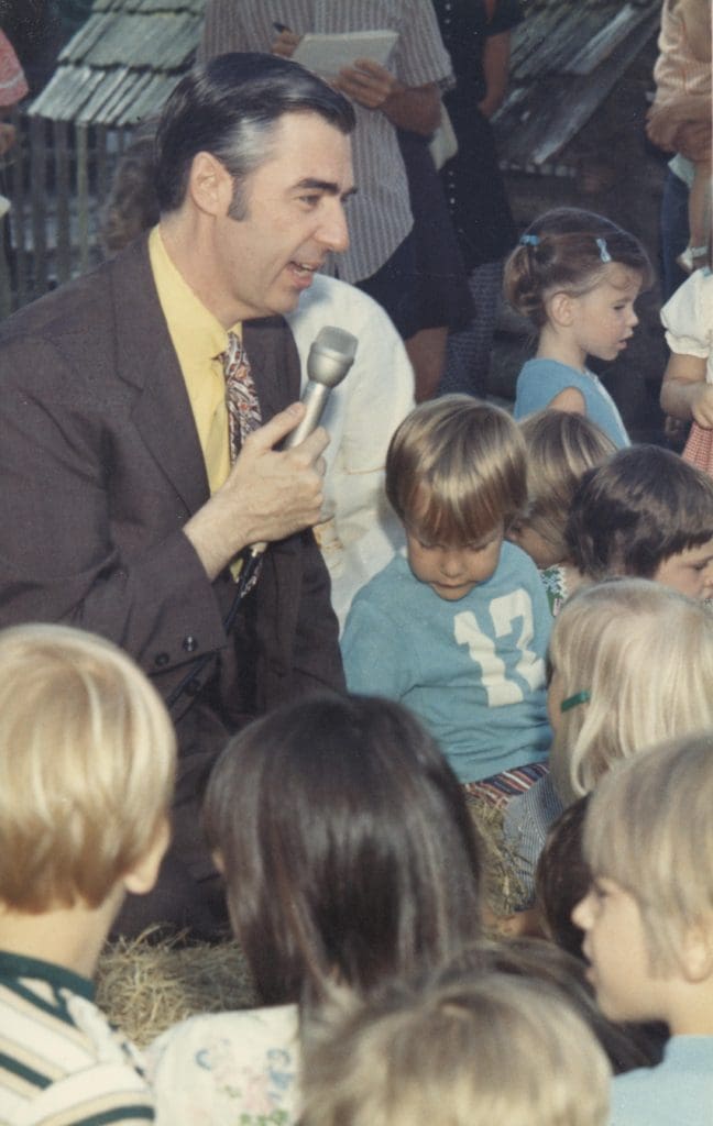 Fred Rogers talks to a group of children. he is holding a microphone